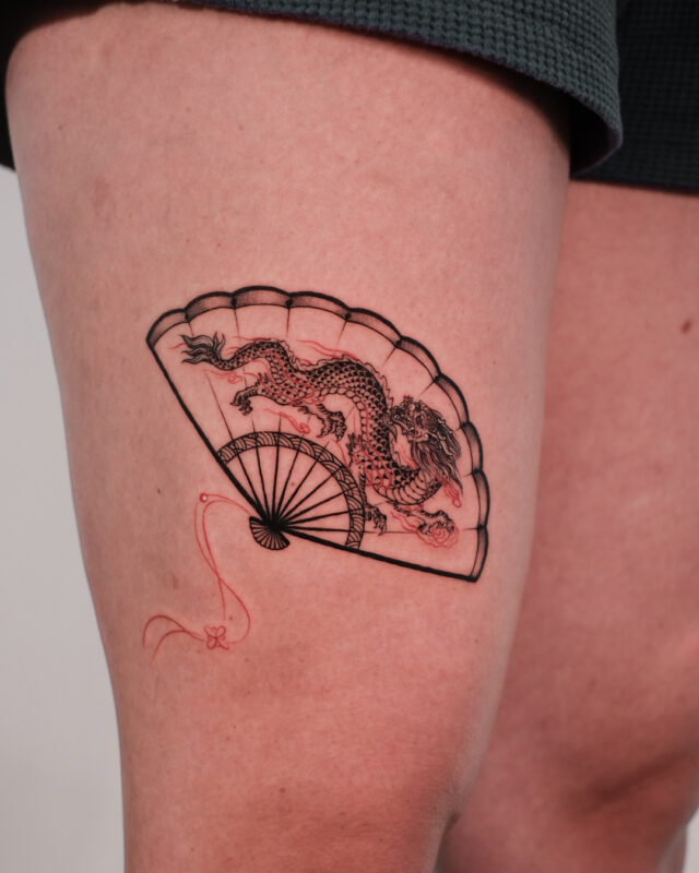 Fine line dragon and folding fan tattoo with red ink details on the thigh by Azone Tattoo Studio in Hanoi