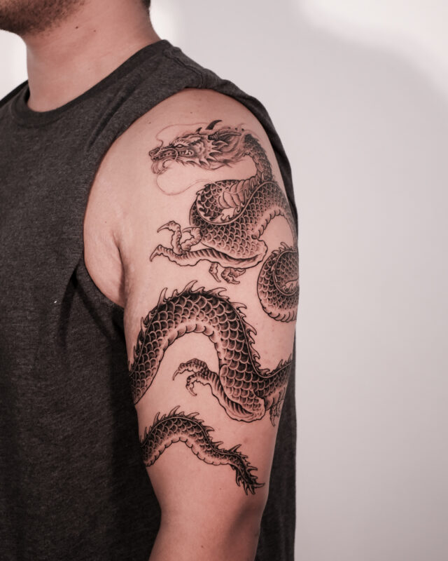 Black and grey dragon tattoo wrapping around the upper arm at Azone Tattoo Studio