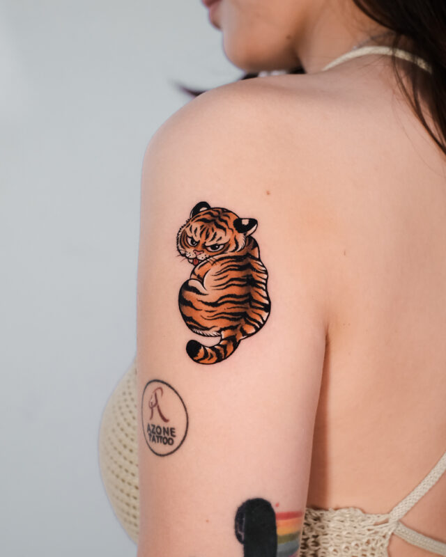 Cute orange and black tiger tattoo with bold outlines and illustrative details on the upper arm.
